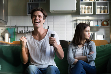 Angry girlfriend sulking while excited boyfriend football fan watching sport game holding remote...