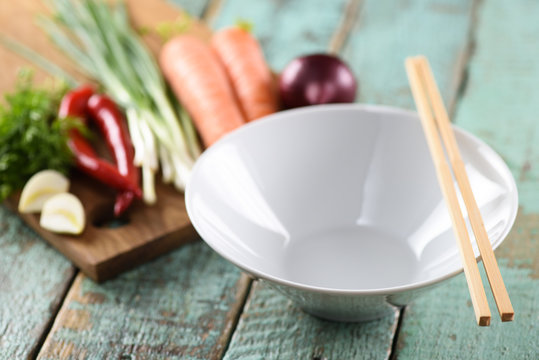 Empty white bowl and raw vegetables, ingredients for Asian udon noodles with chop sticks on wooden blue background