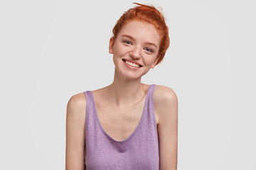 Merry charming foxy female has red hair combed in bun, laughs happily at camera, enjoys free time alone, dressed in loose purple t shirt, stands against white background. People and positiveness