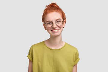 Photo of delighted freckled female with foxy hair tied in knot, has sincere smile, wears glasses and green t shirt, stands over white concrete wall. Student glad to recieve graduation congratulation