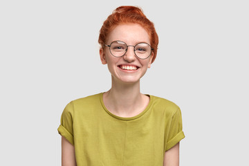 Pretty freckled female with positive smile, wears spectacles and casual loose green t shirt, looks joyfully at camera, satisfied after having picnic with friends, has good day in funny company