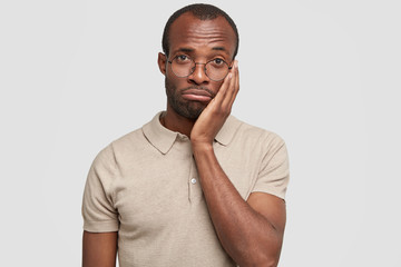 Isolated shot of unhappy African American male purses lips and looks desperately at camera, feels upset after failure, keeps hand on cheek, have some problems, stands against white concerete wall