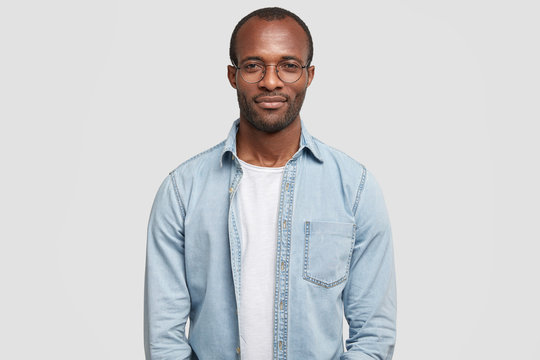 Portrait of confident successful businessman dressed in casual denim shirt, has dark skin, looks self assured, wears big glasses, stands against white background, waits for partners to have meeting