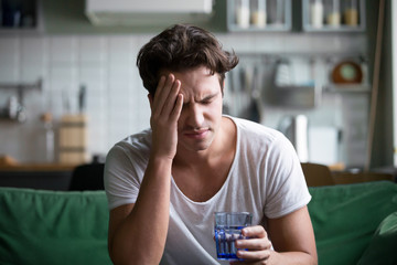 Young man suffering from strong headache or migraine sitting with glass of water in the kitchen, millennial guy feeling intoxication and pain touching aching head, morning after hangover concept - Powered by Adobe