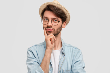 Horizontal shot of confident bearded male keeps finger on chin, has attentive look, dressed in denim shirt, wears straw hat, poses against white background. Self assured man farmer looks seriously