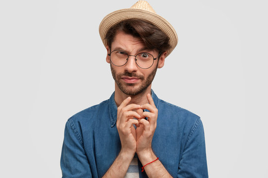 Attractive bearded male keeps hands together and looks with intriguing look, raises eyebrows, has stubble, wears spectacle and starw hat, poses against white background. People and facial expressions