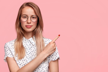 Studio portrait of attractive female student with clever facial expression, wears round spectacles, indicates at right upper corner, has serious look, shows blank space for your advertisement