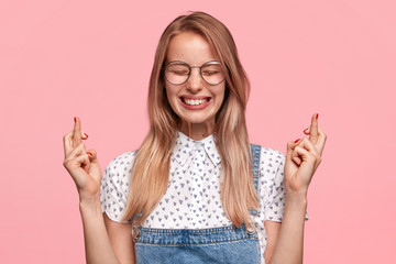 Isolated shot of beautiful female with glad and hopeful expression, crosses fingers and smiles...