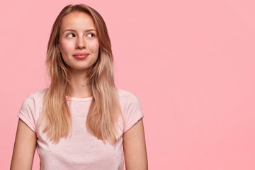 Beautiful cute young female with pensive look, concentrated upwards, has dreamy positive expression, thinks about something pleasant and unforgettable, poses against pink background. Copy space