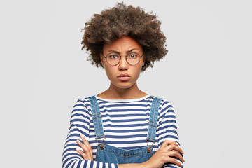 Angry serious African American female student keeps arms folded, frowns face, has mournful expression, looks confidenlty, wears striped sweater, stands against white concrete wall. Facial expressions