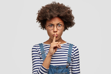 People, ethnicity, conspiracy and body language concept. Pretty annoyed woman with dark skin shows silence sign, demands be quiet and not make noise, raises eyebrows with anger and irritation