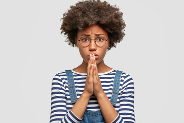Unhappy despondent female asks for apologize and forgiveness, has miserable expression, makes pleading gesture, hopes for something good, stands against white background. Young woman begs indoor.