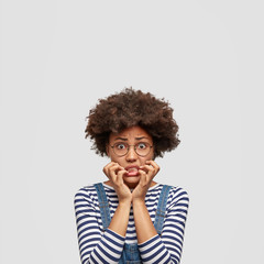 Frustrated dark skinned mixed race female bites finger nails nervously, being in panic as hears bad news, worries before perfomance on stage, poses against white background with blank copy space