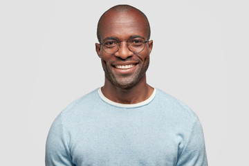 Middle aged cheerful dark skinned male with shining smile, wears light blue sweater, round spectacles, achieves everything by himself, stands against white studio wall, has relaxed face expression - obrazy, fototapety, plakaty