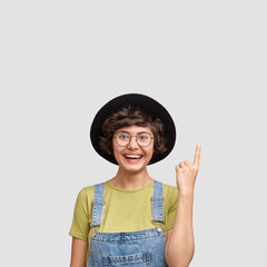 Hey, look there! Beautiful young Caucasian female with broad smile, indicates upwards with fore finger, wears denim overalls and black hat, poses against white background, blank space for promotion