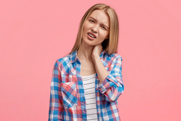 Frustrated displeased blonde young cute female has strong pain in neck, tries to make massage, makes pain grimace, feels overworked and tired, wears casual checkered shirt, stands indoor alone.