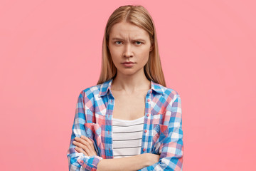 Studio shot of beautiful young female has sullen expression, frowns face in discontent and keeps hands crossed, wears checkered shirt, being offended by someone, isolated over pink background