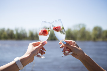 Two female hands holding a glass of champagne with strawberries. Background river