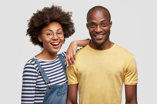 Happy dark skinned female with gentle smile, leans at best friend`s shoulder, have fun together, wear casual clothes and spectacles, pose together against white background. African American family