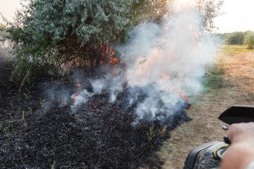 Forest wildfire. Burning field of dry grass and trees. Heavy smoke against blue sky. Wild fire due to hot windy weather in summer. rescuer inspecting disaster area by atv quad bike