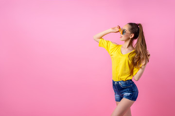 Female teenager in sunglasses and summer clothes standing in studio with flying ponytail on pink background - obrazy, fototapety, plakaty
