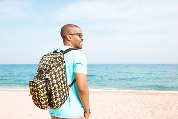 Portrait of attractive young African-American hipster wearing t-shirt and sunglasses walking at summer day on the beach with backpack