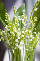 A bouquet of white flowers from lilies of the valley with green leaves standing in a glass on the windowsill