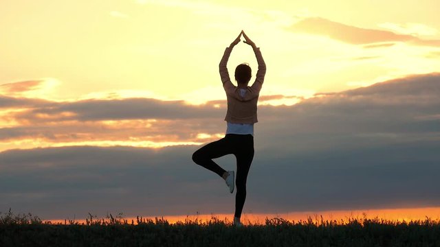 Young woman silhouette practicing yoga on the meadow at sunset. Girl with a slender figure practices yoga at sunset. Sport and health.