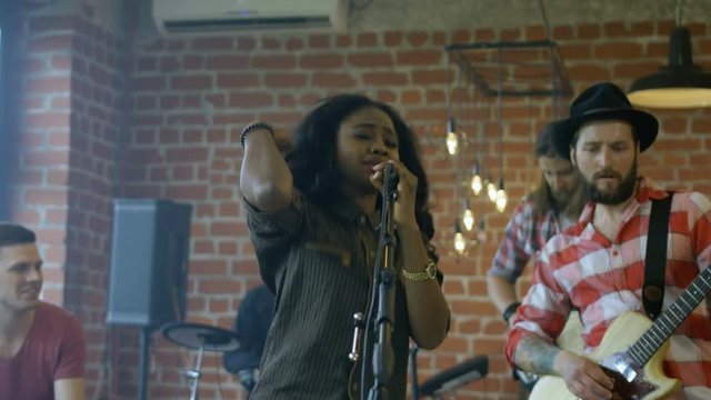 Stylish African-American woman singing into microphone having performance with rock band in studio