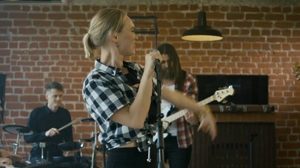 Young casual woman singing with emotions on stage having fun with musicians playing instruments on rehearsal