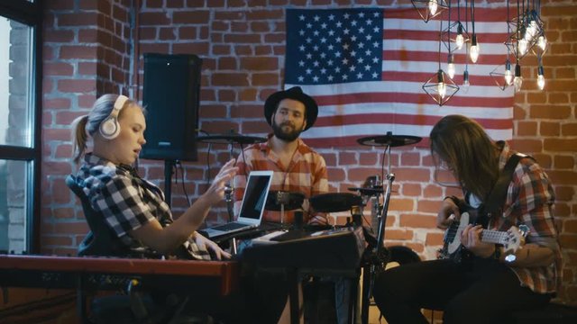 Cheerful blonde girl at synthesizer and men on drums and bass guitar creating music together in studio with American flag