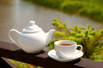 Tea on the wooden railing on the river bank. A cup of tea and a teapot against the background of a river and green grass. Tea and summer bright sun.