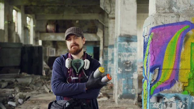 Portrait of bearded guy graffiti painter standing in old empty building with graffiti painting in background and holding spray-paint. Man has gas mask and wearing gloves.