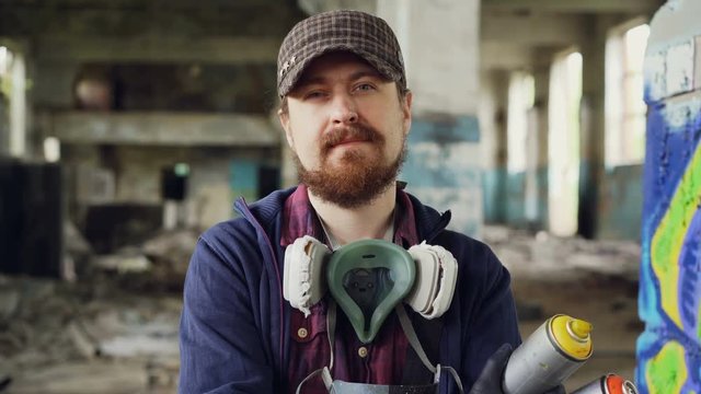 Close-up portrait of handsome bearded man graffiti artist standing inside abandoned building wearing cap, gloves and pespirator and holding spray paint