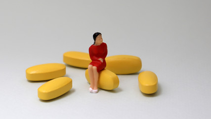A miniature woman sitting on a pills beside several yellow pills.