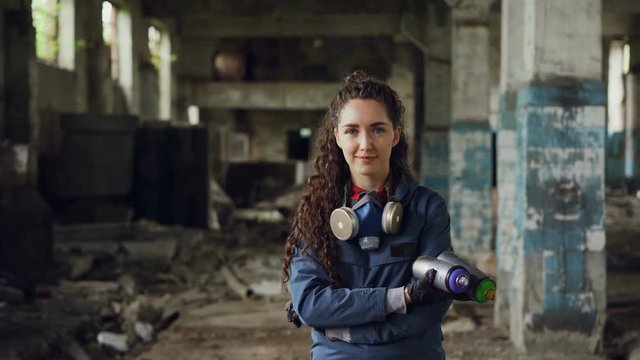Portrait of smiling beautiful girl graffiti painter standing in old abandoned building, holding spray paints and looking at camera. Woman has protective mask and gloves.