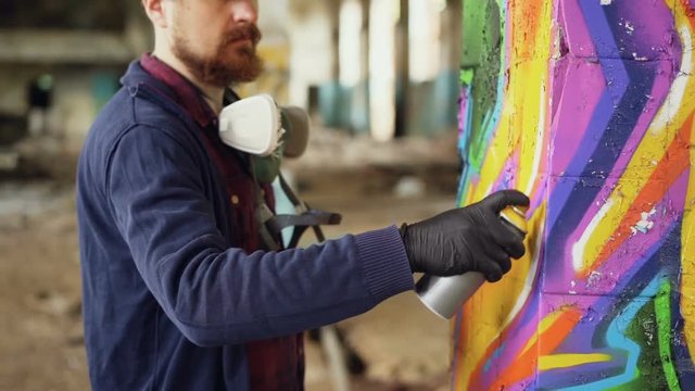 Bearded man graffiti painter is using aerosol paint to decorate pillar in old industrial building. Modern urban art, creative young people and hobby concept.