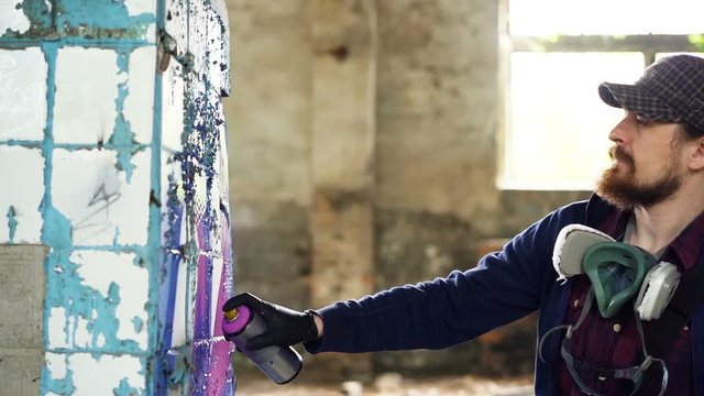 Attractive bearded guy is holding aerosol paint and drawing graffiti on pillar inside spacious abandoned house. Creative people, empty buildings and modern art concept.