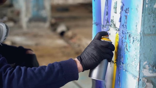 Close-up shot of male hand in protective glove holding aerosol paint and painting graffiti on pillar inside abandoned building. Modern art, abstract images and people concept.