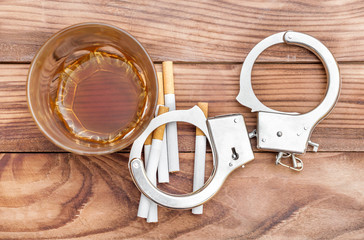 Glass of alcohol drink with cigarettes and handcuffs on wooden table. Bad dependence and habits....