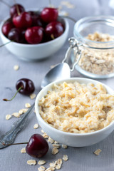 Diet and healthy Breakfast: oatmeal with cherries in a white plate on a gray tablecloth, in the background dish with cherries and a can of oatmeal