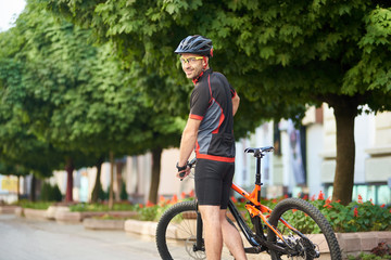 Athletic man cyclist smiling looking over shoulder posing in cycling garment and protective helmet near bike next to green trees in city street. Concept of healthy life, outdoor activities
