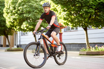 Obraz premium Athletic guy cyclist in cycling clothes and protective helmet, riding bike down empty city center alley surrounded by green trees. Sportsman training outdoors improving skills.