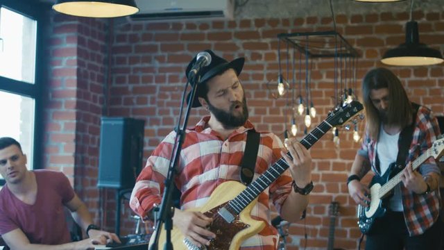 Casual hipster man with beard playing guitar and singing expressively while playing music with band in studio