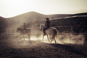 ride horses lessons at the sunset with golden backlight. dust and two horses and caucasian cowboy style people. vacation and leisure outdoor in feeling with the nature and the animals