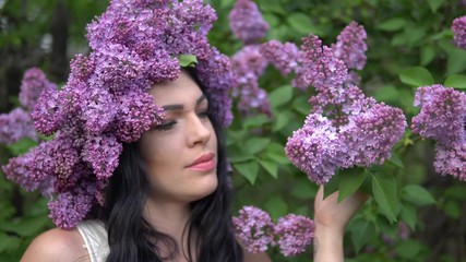 Beautiful brunette girl with a wreath on her head admiring and sniffing lilac flowers