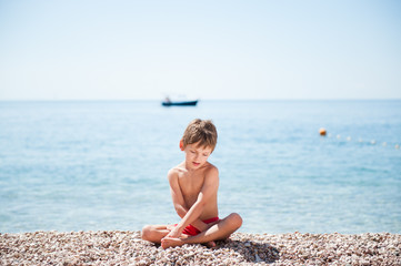 Fototapeta na wymiar handsome small kid sitting on sea shore beach with sea horizon with boat behind