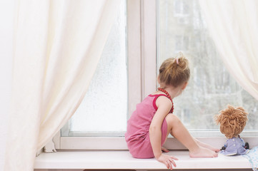 Sad little girl with a doll waiting for her mother near the window