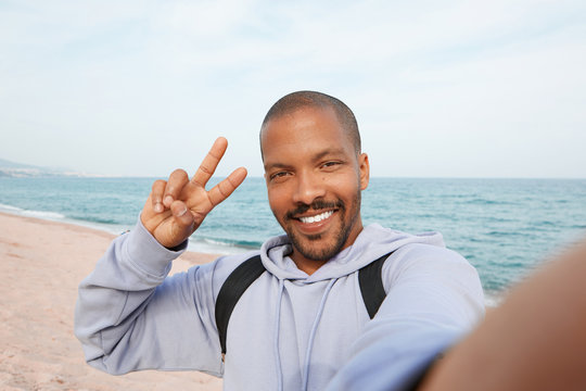 Happy young African-American hipster man taking selfie and making hand victory symbol on the beach. Cloudy sky on blurred background