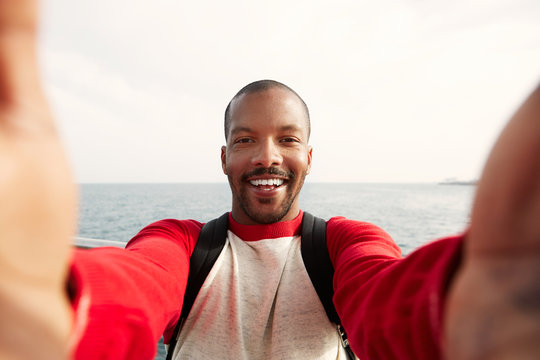 Handsome smiling young African-American hipster man taking two hands selfie with happy face outdoors of city. Blue sea on the blurred background